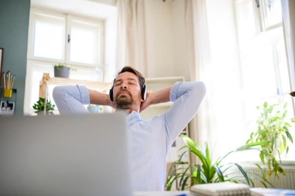 Tired mature businessman with headphones and laptop indoors in home office, resting.