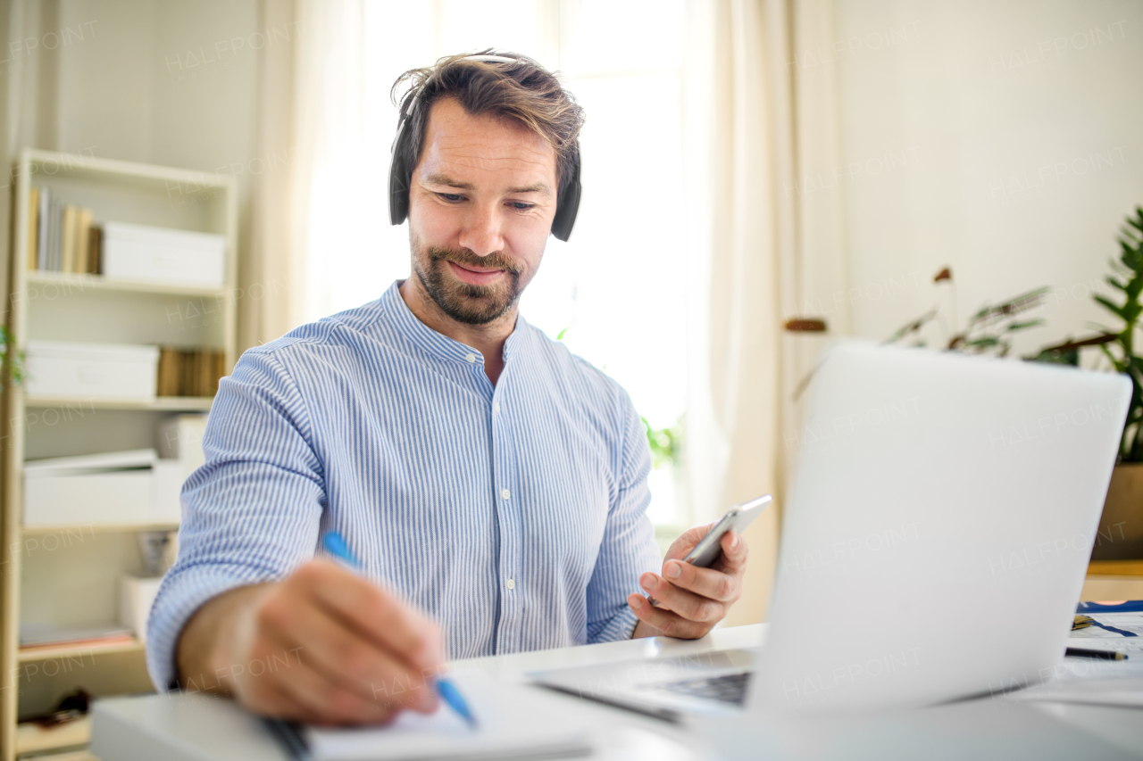 Front view of mature businessman with headphones and laptop indoors in home office, working.