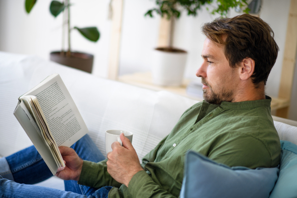 Happy man sitting on sofa and reading book at home, lockdown concept.