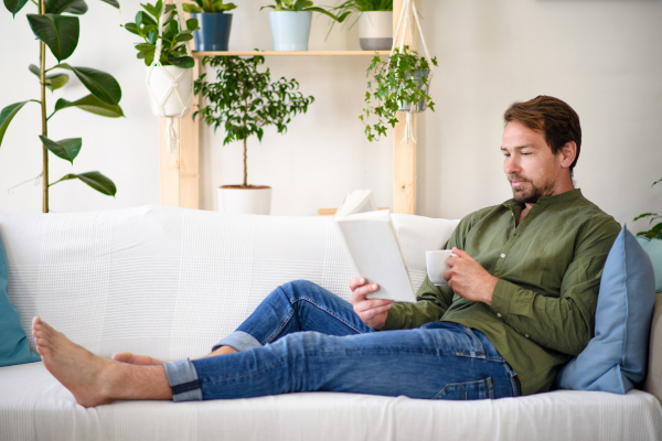 Happy man sitting on sofa and reading book at home, lockdown concept.