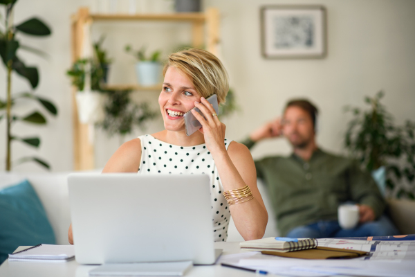 Cheerful young businesswoman with laptop and telephone indoors in home office, working.