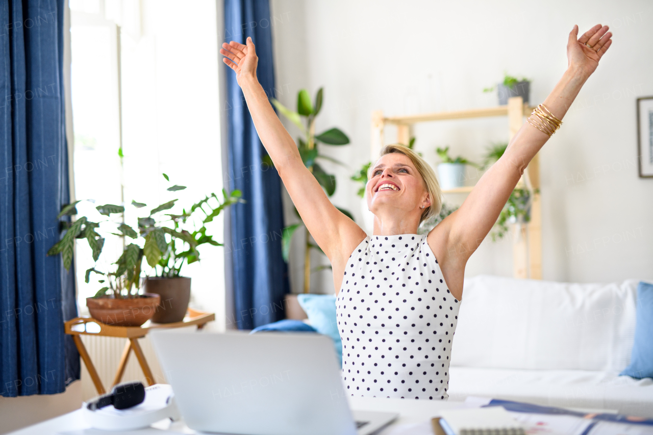 Cheerful young businesswoman with laptop indoors in home office, stretching arms when working.