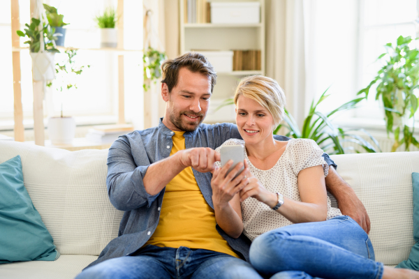 Front view of happy couple in love sitting indoors at home, using smartphone.