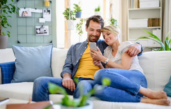 Front view of happy couple in love sitting indoors at home, using smartphone.