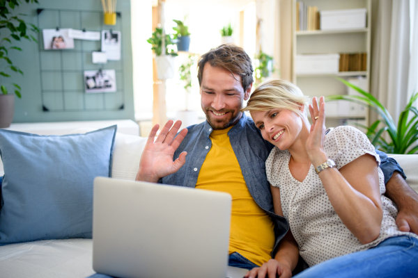 Front view of happy couple having video call on laptop indoors at home, waving.