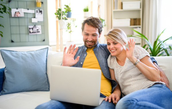 Front view of happy couple having video call on laptop indoors at home, waving.