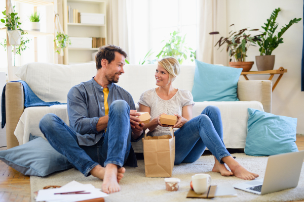 Front view of happy couple sitting on floor indoors at home, eating hamburgers.