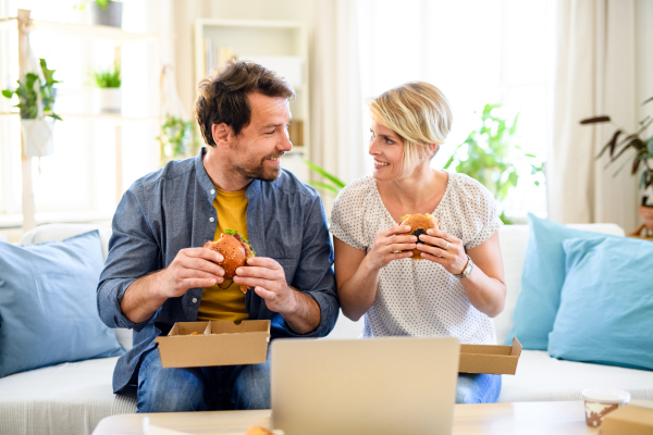 Front view of happy couple sitting on sofa indoors at home, eating hamburgers.