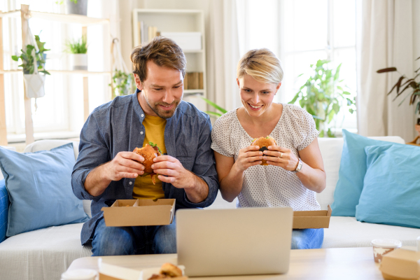 Front view of happy couple sitting on sofa indoors at home, eating hamburgers when watching film.