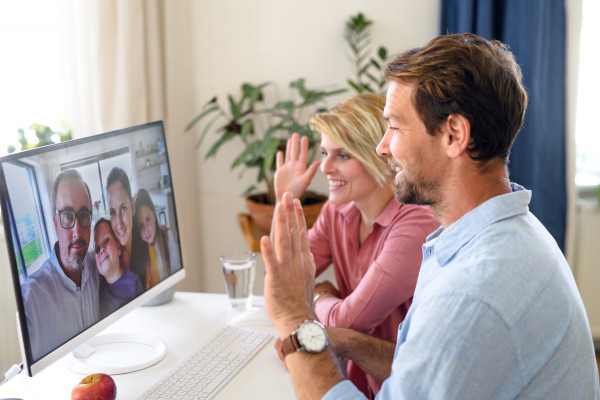 Front view of happy couple having video call with friends on computer indoors at home, waving.