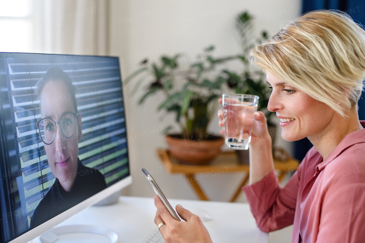 Young businesswoman having video call on computer in home office, using smartphone.