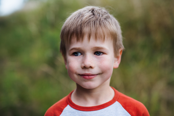 A portrait of blond school boy standing in nature, looking at camera.