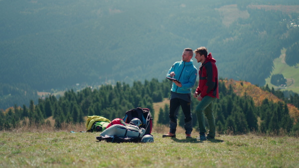 Two paragliders men standing on a top of mountain with tablet and looking at view.