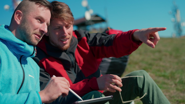 Two paragliders men sitting on the top of the mountain with tablet and looking at view.