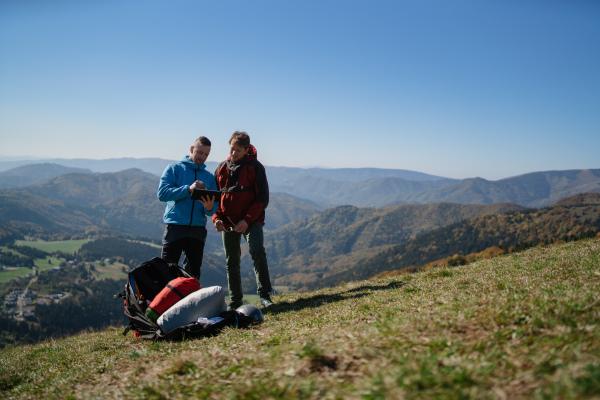 Two paragliders men standing on a top of mountain with tablet and looking at view.