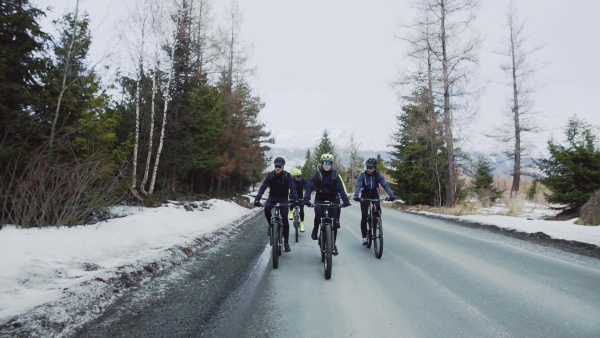 A group of mountain bikers riding on road outdoors in winter, mountains in the background. Slow motion.