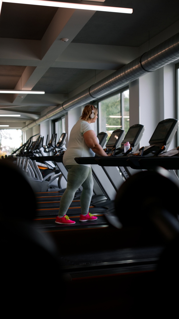 A vertical footage of overweight woman with headphones exercising on treadmill in gym