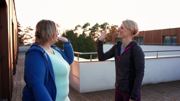 A happy overweight woman high fiving with personal trainer outdoors on gym terrace.