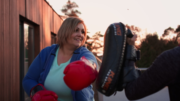 An overweight woman training boxing with personal trainer outdoors on terrace.