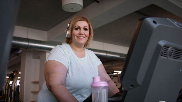 Mid adult overweight woman with headphones running on treadmill in gym