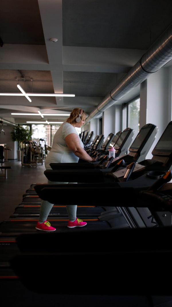 A vertical footage of overweight woman with headphones exercising on treadmill in gym