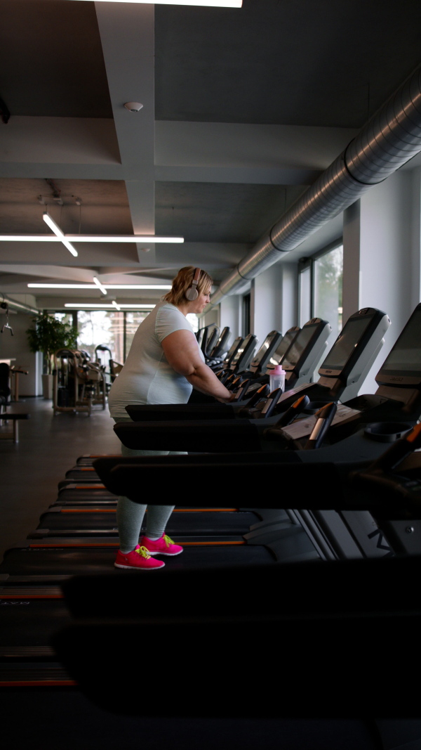 A vertical footage of overweight woman with headphones exercising on treadmill in gym