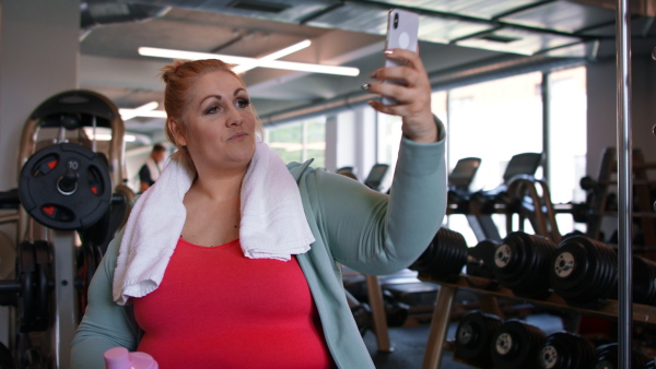 A happy fat woman after exercise indoors in gym, taking selfie.