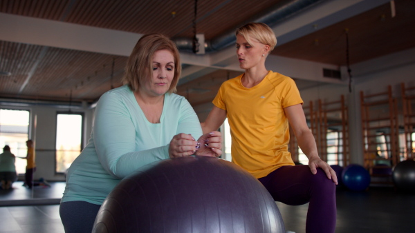 A happy overweight woman exercising with personal trainer on fintess ball in gym