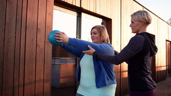 An overweight woman holding ball and exercising with personal trainer in outdoors on gym terrace.