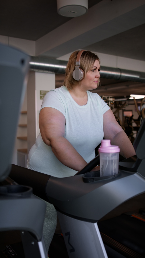 Mid adult overweight woman with headphones running on treadmill in gym