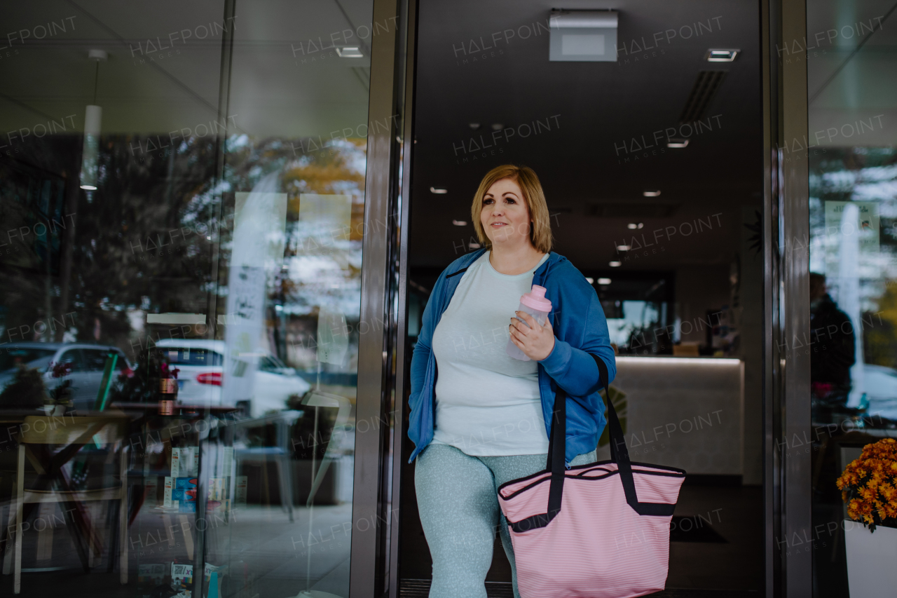 A happy overweight woman in sports clothes coming out from gym.