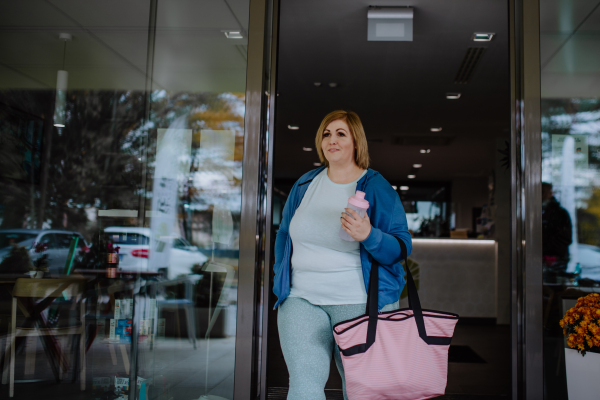 A happy overweight woman in sports clothes coming out from gym.