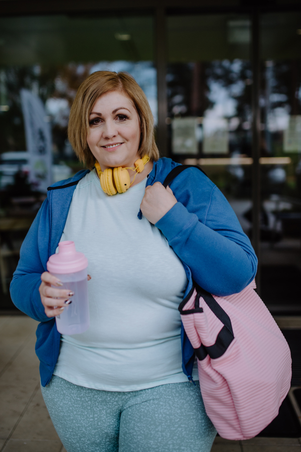 A happy overweight woman in sports clothes looking at camera outdoors in front of fitness center