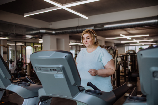 Mid adult overweight woman with headphones running on treadmill in gym