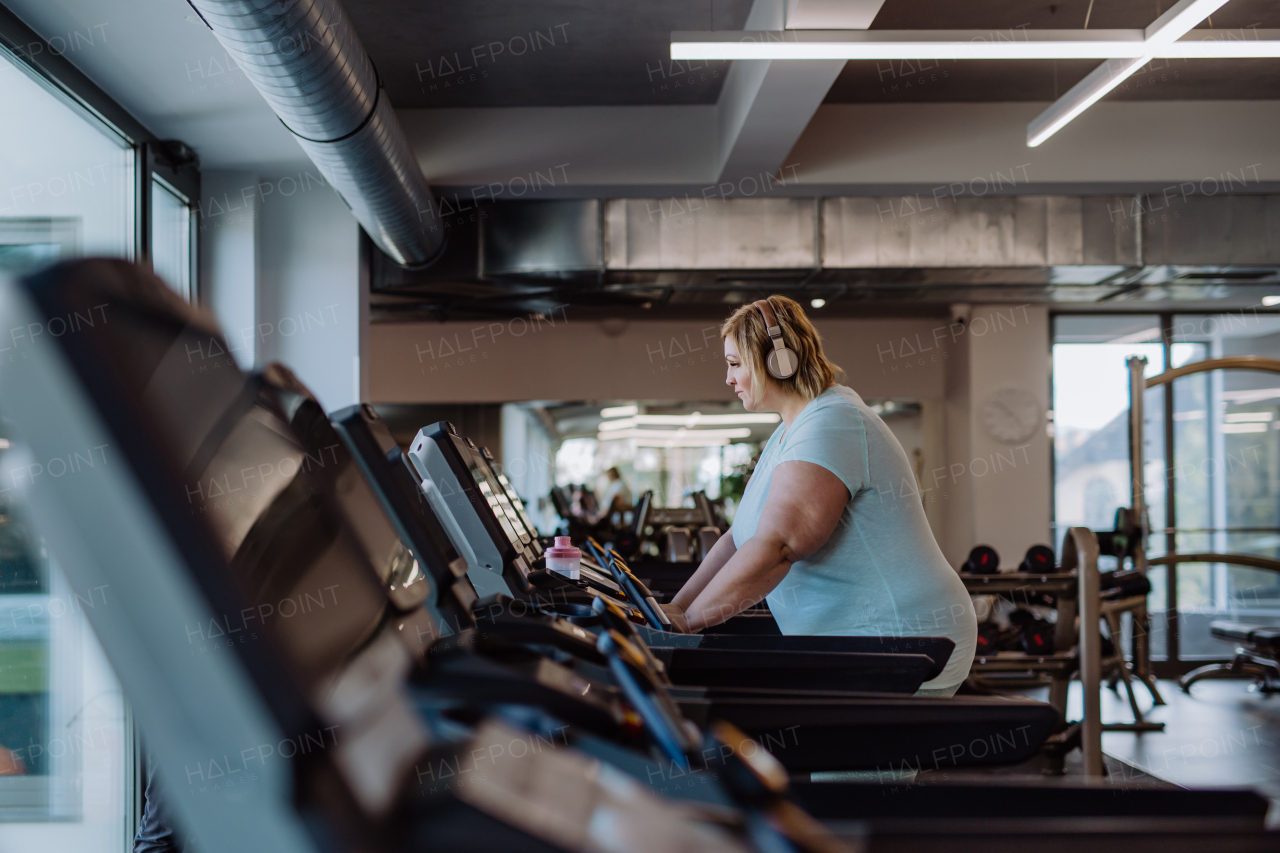 Mid adult overweight woman with headphones running on treadmill in gym