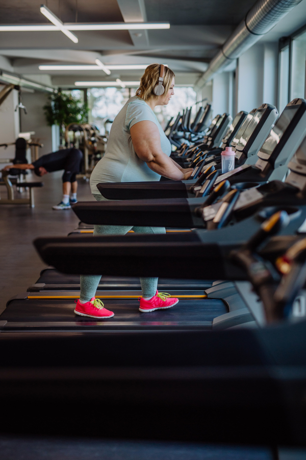 Mid adult overweight woman with headphones running on treadmill in gym