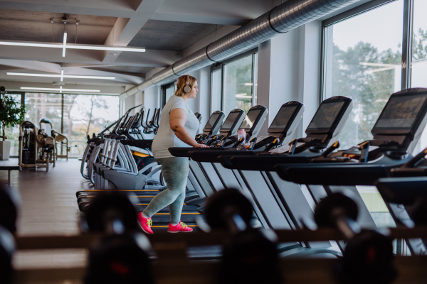 Mid adult overweight woman with headphones running on treadmill in gym