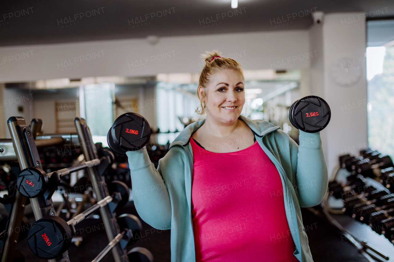 A plus size woman training and lifting dumbbells indoors in gym, looking at camera