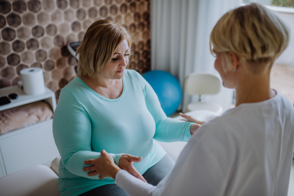 A mid adult physiotherapist woman exercising with overweight woman indoors in rehabilitation center