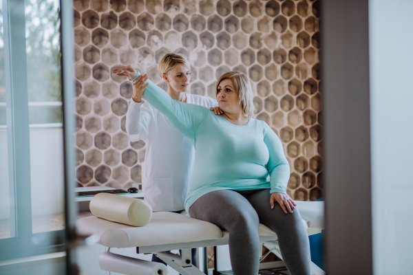 A mid adult physiotherapist woman exercising with overweight woman indoors in rehabilitation center