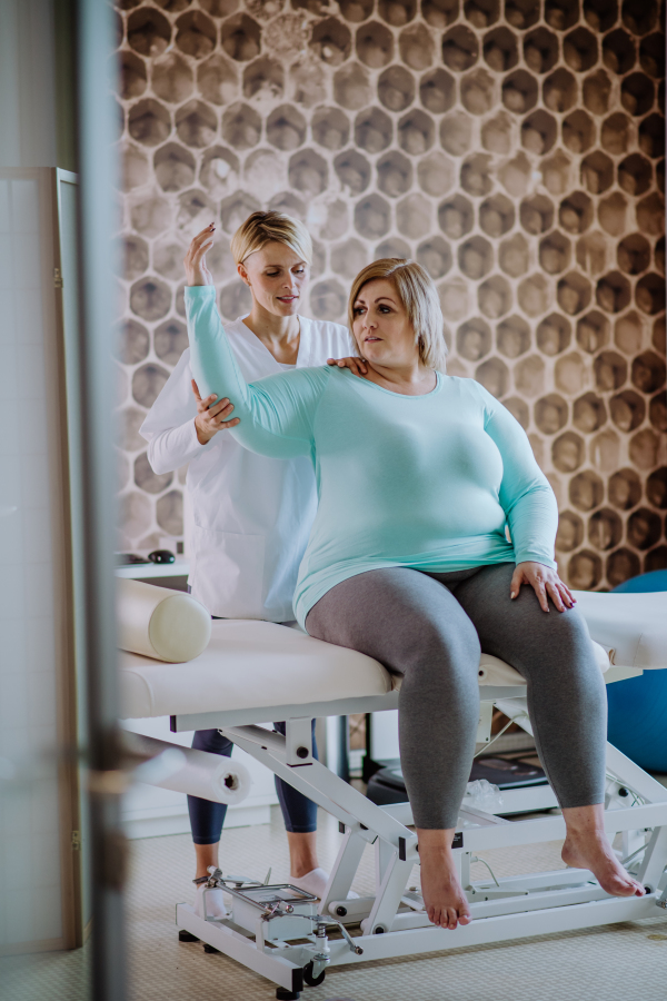 A mid adult physiotherapist woman exercising with overweight woman indoors in rehabilitation center