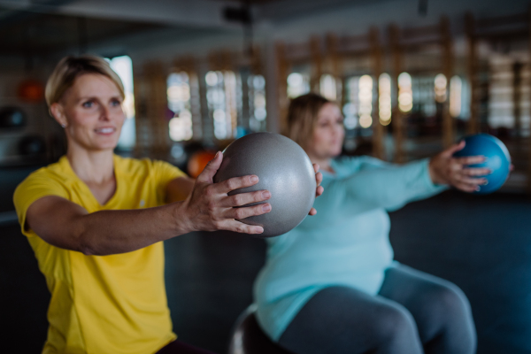 A personal trainer training overweight woman, sitting on fintess bal and exercising in gym.