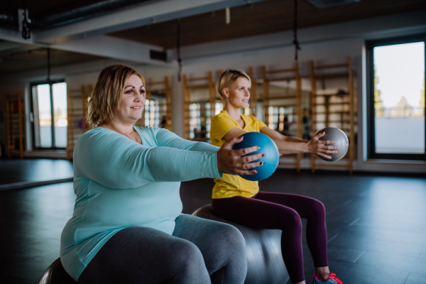 A happy overweight woman sitting on fintess ball and exercising with personal trainer in gym