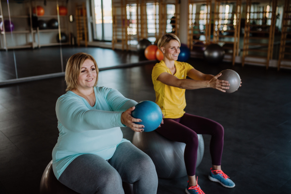 A happy overweight woman sitting on fintess ball and exercising with personal trainer in gym