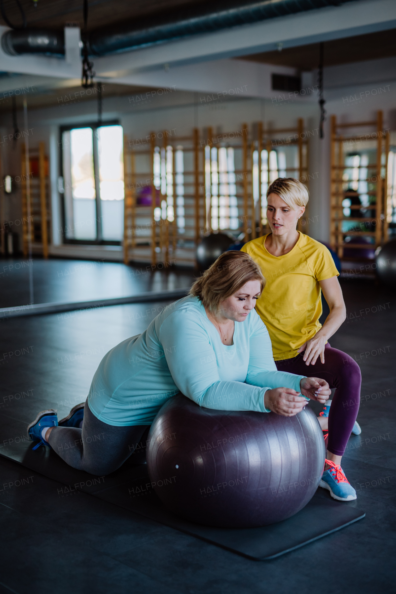 An overweight woman exercising with personal trainer on fintess ball in gym