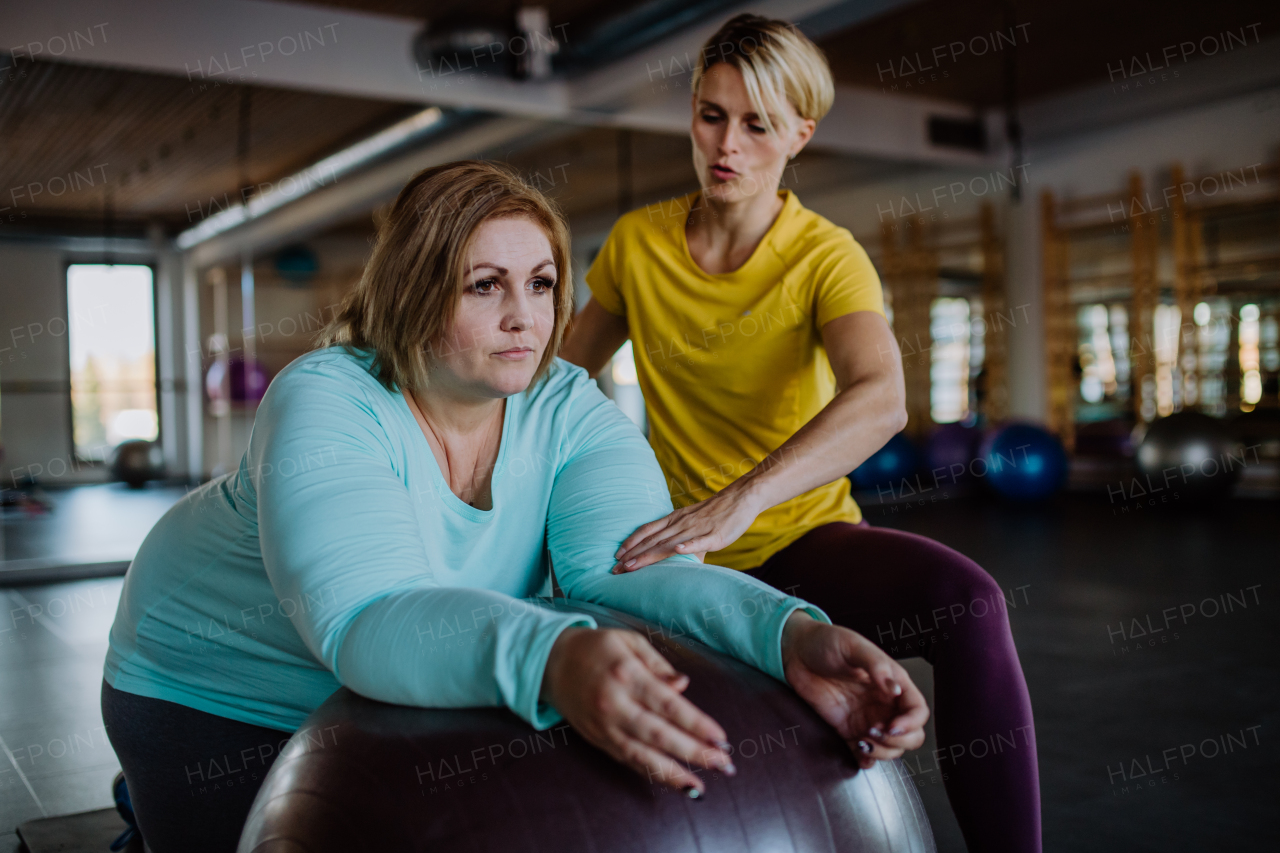 A happy overweight woman exercising with personal trainer on fintess ball in gym