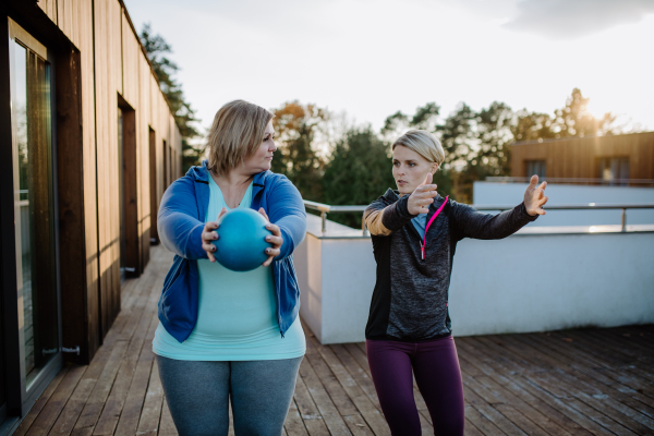 An overweight woman holding ball and exercising with personal trainer in outdoors on gym terrace.