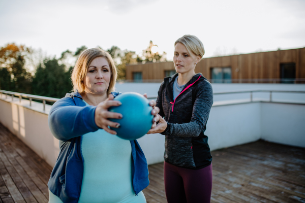 An overweight woman holding ball and exercising with personal trainer in outdoors on gym terrace.