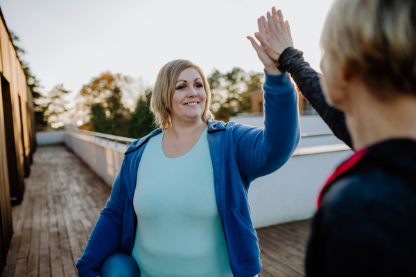 A happy overweight woman high fiving with personal trainer outdoors on gym terrace.