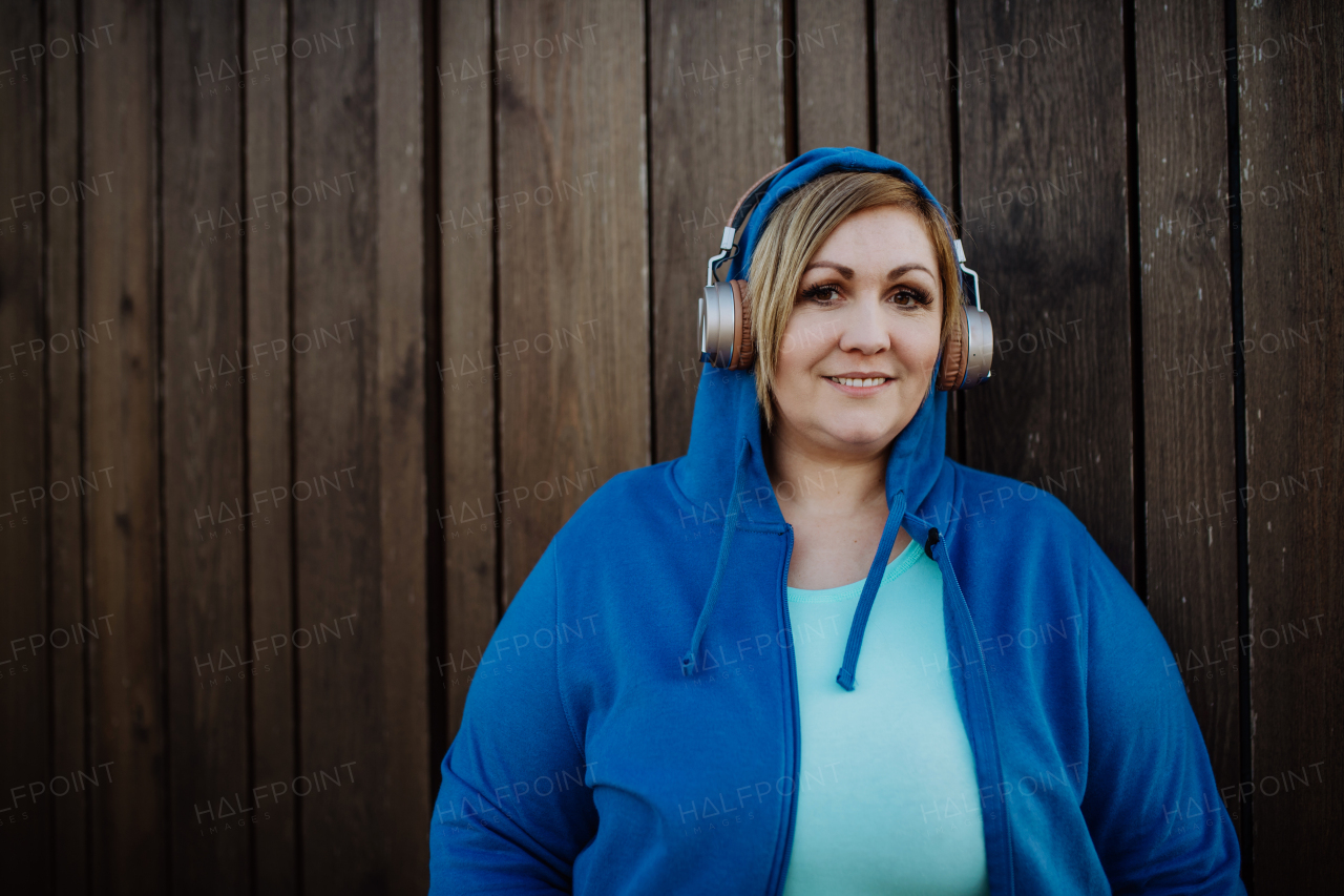 A happy overweight woman in sports clothes with headphones looking at camera against wooden wall